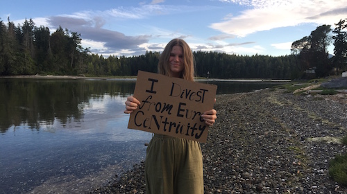 A young girl stands in front of a river holding a sign that says "I divest from Euro Centrism"