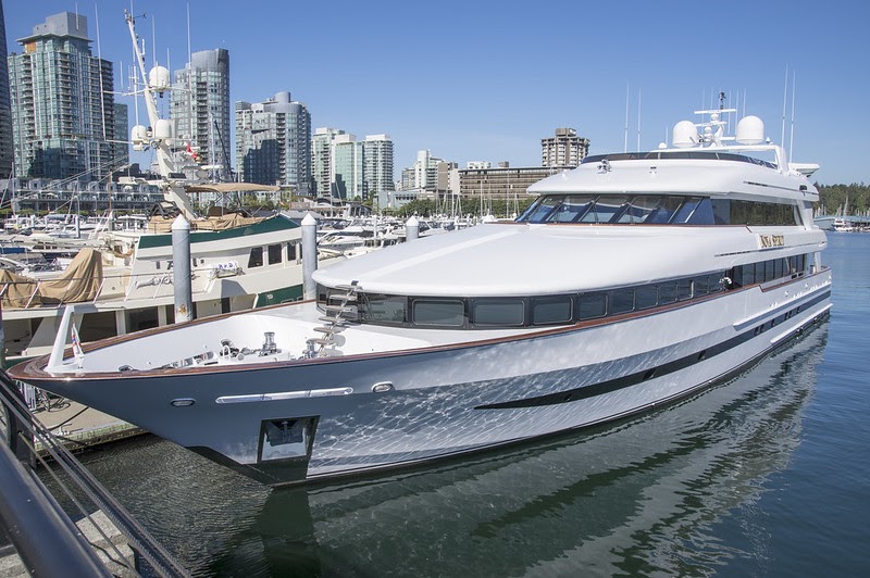Looking at a 147 foot yacht from its port side bow, tied up at a marina with a city scape in the background.