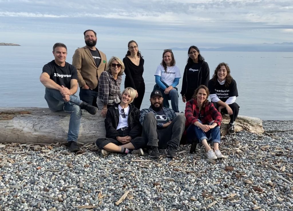 A group of ten people smiling for the camera, either sitting and standing in a group around a log on a pebble beach with ocean in the background.