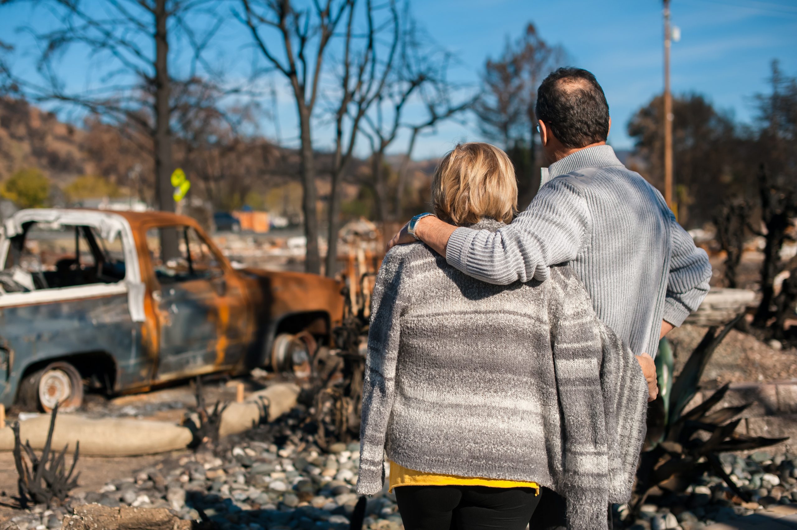 A man and woman standing together looking away from camera, with the man's arm over the woman's shoulder, as they look at a fire-damaged property including a burnt tree and pick up truck.