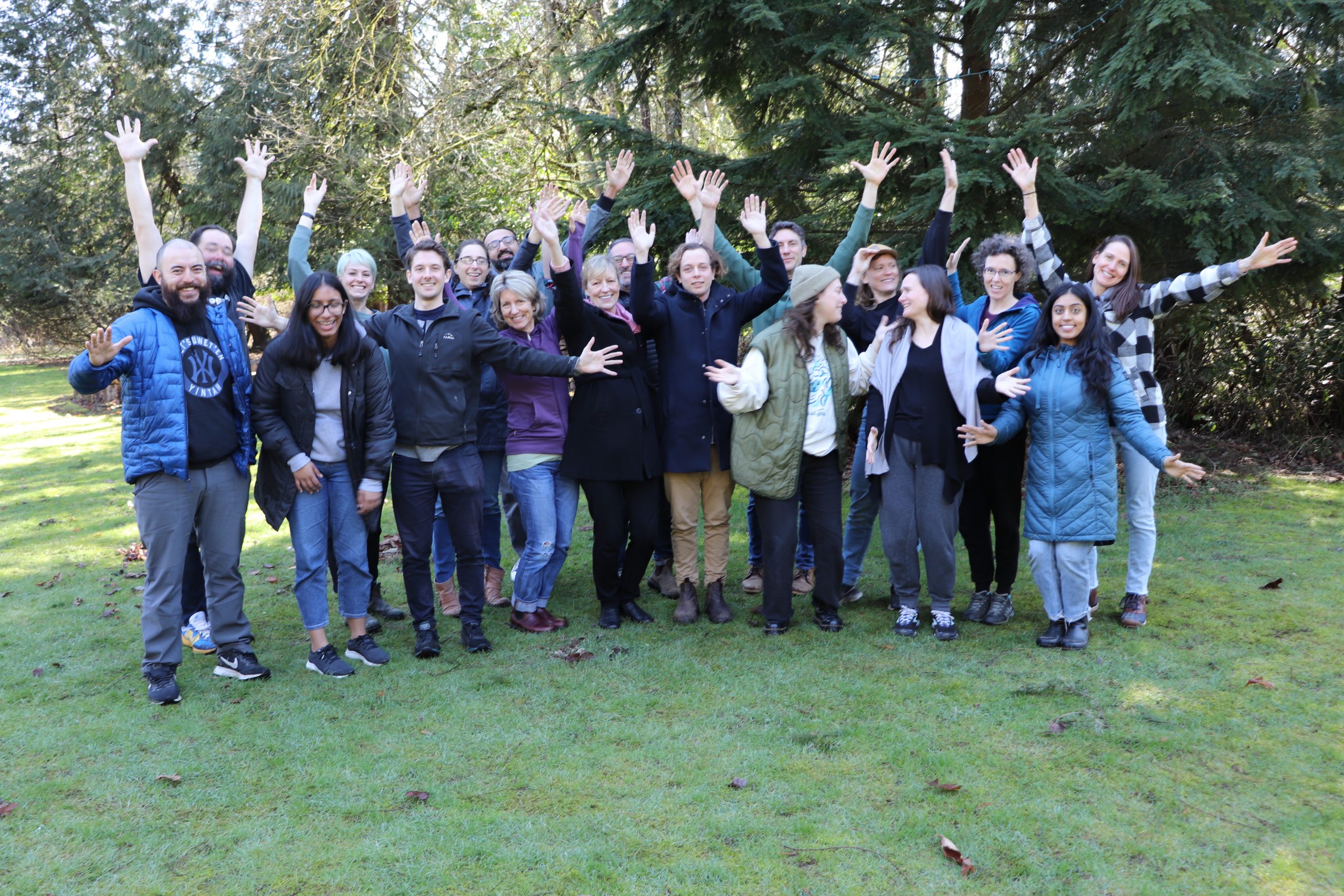 Group of about 20 people standing together on a lawn with their hands in the air and smiling for the camera with trees in the background.