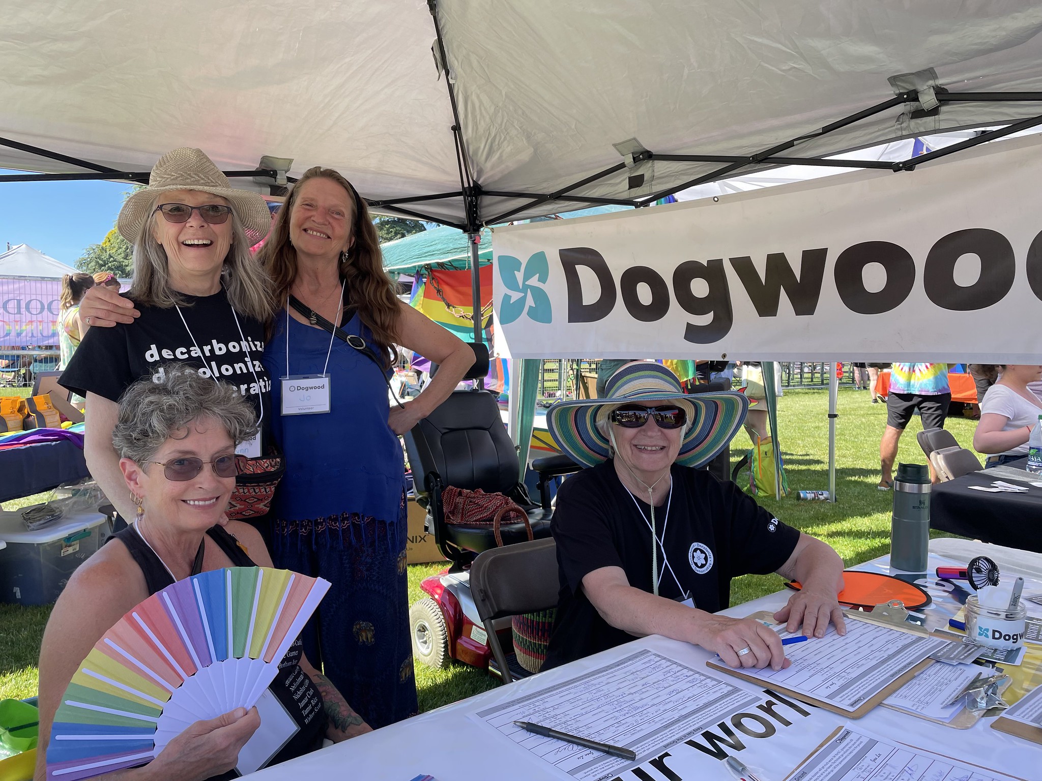 Four people smiling for the camera, two sitting and two standing, behind a table with petition clipboards all across and a banner that says "Dogwood" hanging from the pop up tent overtop of them all.